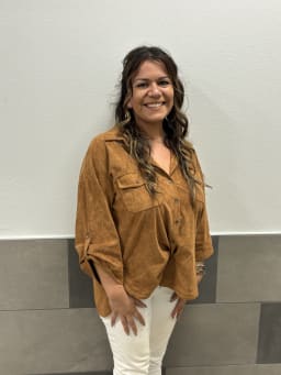 Brown skin female wearing brown long sleeve shirt, long hair, smiling against a white wall.