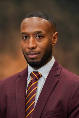 A well-dressed man with a beard wearing a burgundy suit and a striped tie stands in front of a blurred background.