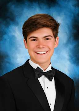 A young man in a black tuxedo with a bow tie is smiling against a blue background with some smoke or haze.