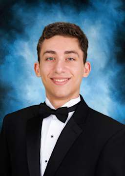 A young man in a black tuxedo with a bow tie is smiling against a blue background with some smoke or haze.