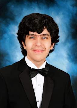 A young man wearing a black tuxedo with a bow tie stands in front of a blue background, smiling confidently.
