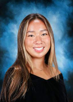 A smiling young woman with long brown hair stands against a blue and smoky background.