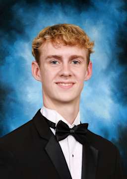 A young man with blond, curly hair is smiling while wearing a black tuxedo against a blue, smoky background.