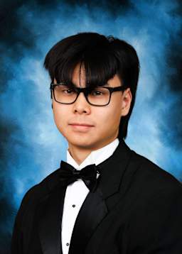 A young man wearing a formal black suit and bowtie stands against a blue smoky background, his expression serious and focused.