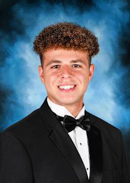 A young man with curly brown hair and a warm smile, dressed in a black tuxedo, stands against a blue smoky background.