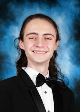 A smiling young person wearing a formal black suit and bowtie stands against a blue background.
