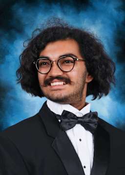 A smiling man with curly hair and glasses, wearing a tuxedo, stands against a vibrant blue background.