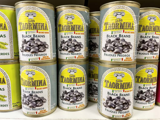 Cans of Taormina black beans are stacked on a shelf. The labels display images of beans and text in English and Spanish. Bright and colorful packaging.