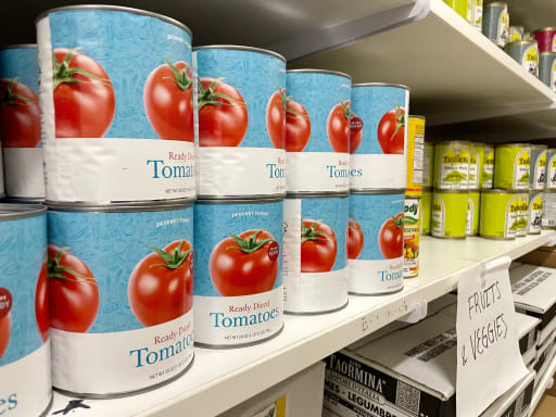 Cans of tomatoes are neatly stacked on shelves beside other canned goods under a sign labeled "Fruits & Veggies," conveying a tidy pantry scene.
