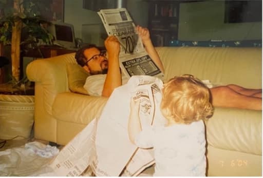 A man reading a newspaper on the couch while a girl looks up at him