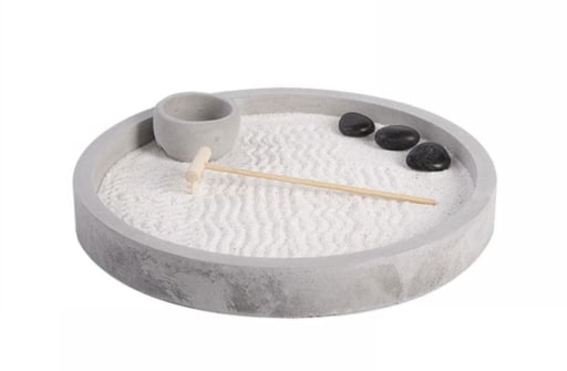 A round tabletop Zen garden features white sand with delicate raked patterns, three black stones, and a small wooden rake. The tone is calm and minimalist.
