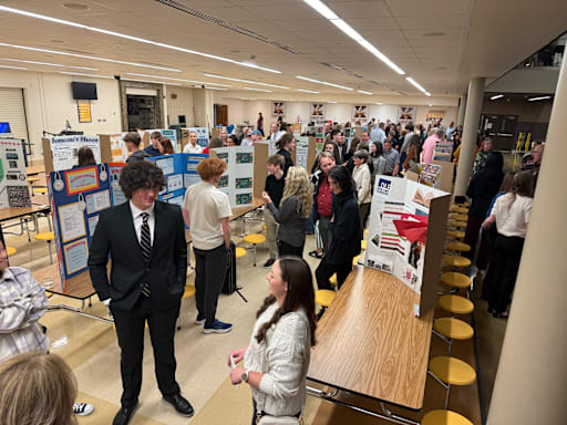 People mill between cafeteria tables with tri-fold cardboard presentations on them.