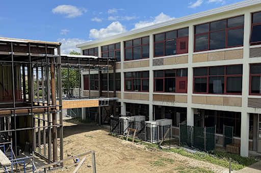 A construction site with a steel frame in the foreground and a building with many windows in the bac…