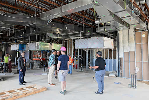 Several construction workers in hard hats stand in a large, unfinished room with exposed ductwork an…