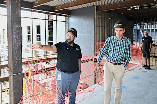 Two men in hard hats stand in the foreground of a construction site, with unfinished walls and scaff…