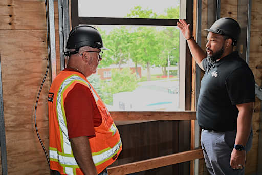 Two construction workers in hard hats and safety vests discuss a window frame inside a building unde…