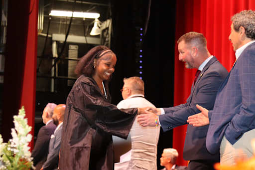 A woman in a black robe shakes hands with a man in a suit on a stage with red curtains in the backgr…
