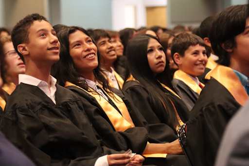 A group of young people in graduation gowns smile and look upwards in a crowded auditorium.