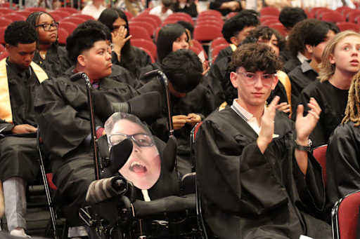 A group of graduates in black gowns sit in red chairs, with a cutout of a smiling face in glasses in…