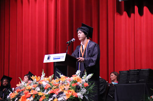 A young man in a graduation cap and gown speaks at a podium in front of a vibrant orange and white f…