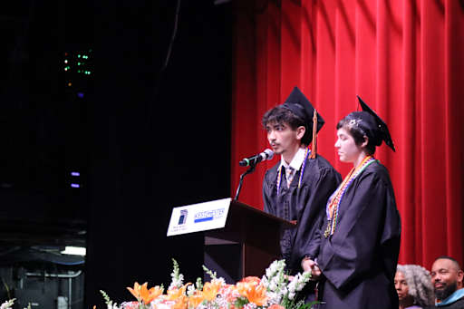 Two graduates in caps and gowns stand at a podium with a microphone in front of a red curtain, while…