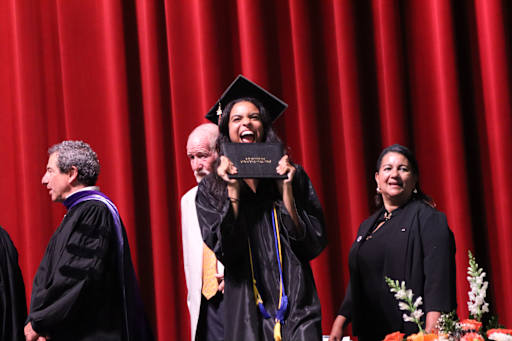 A jubilant graduate in a cap and gown holds up her diploma in front of a red curtain backdrop.