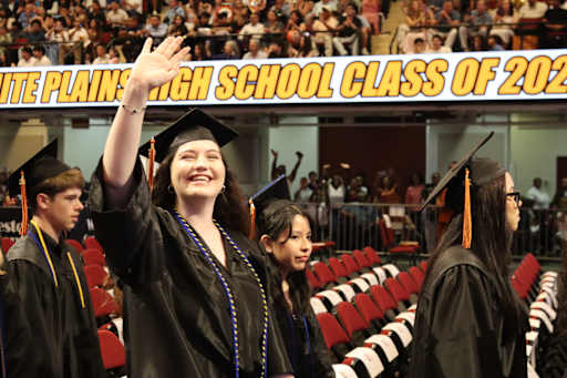 A smiling graduate in the foreground waves to the crowd in the background, which is filled with spec…