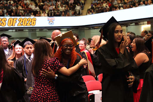 Graduates in caps and gowns embrace in the foreground, while a cheering crowd fills the stadium seat…