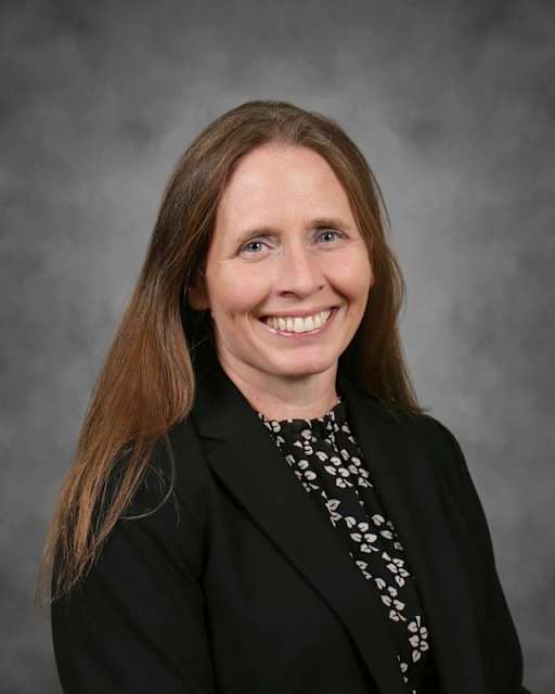 A smiling woman with long brown hair, wearing a black blazer over a patterned shirt, is set against a plain gray background.