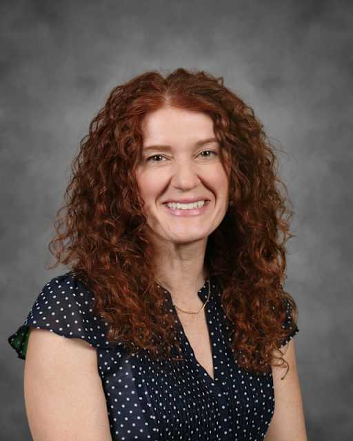 A smiling woman with long, curly red hair and a polka-dot shirt is set against a gray, textured background.