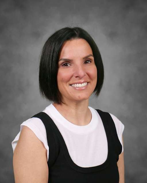 A smiling woman with dark hair and a white shirt under a black vest is posed against a textured gray background.