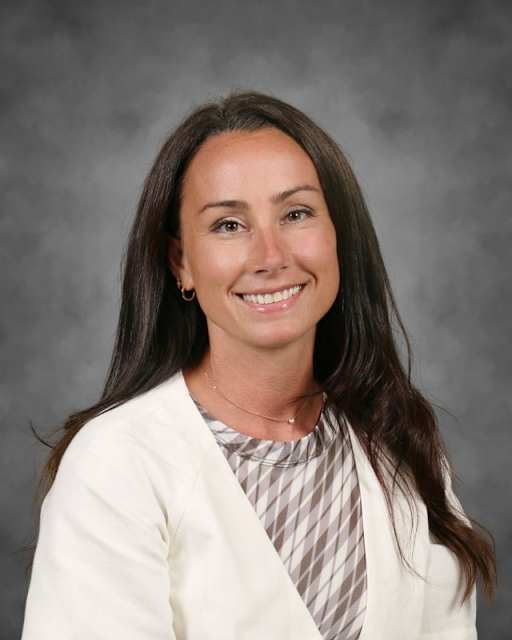 A woman with long dark hair and a white jacket smiles against a gray, textured background.