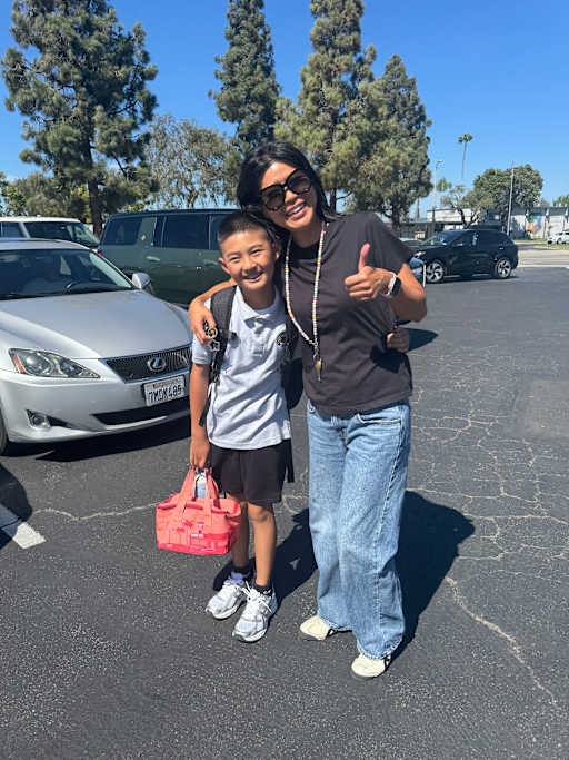 A smiling boy with a backpack and lunchbox stands next to a woman giving a thumbs-up, with cars and trees in the background.
