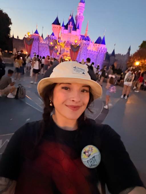 A smiling woman in a bucket hat and Disney-themed attire poses for a selfie in front of a brightly lit Sleeping Beauty Castle at dusk, with a crowd of people in the background.