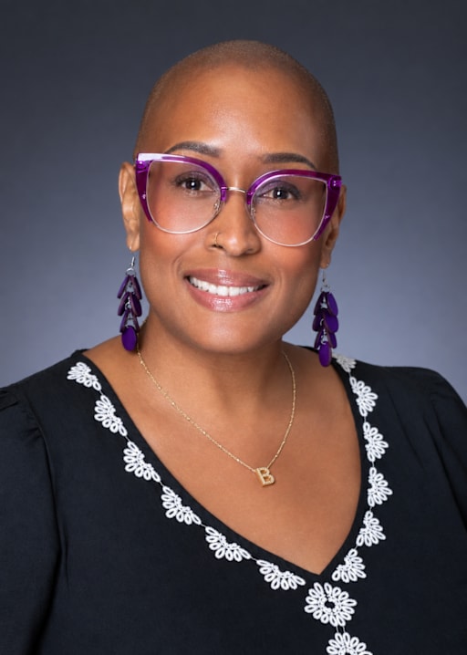 A bald woman wearing purple glasses, purple earrings, and a black embroidered top poses against a dark gray background.