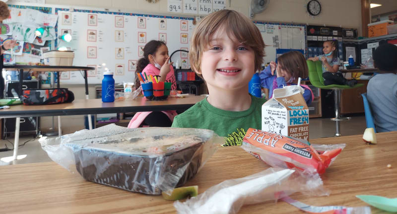 Young student smiles while eating lunch at desk