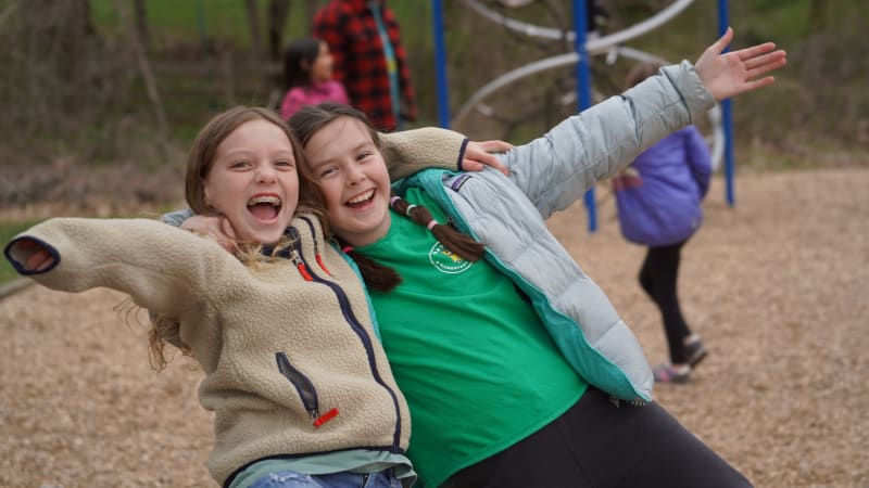 Two girls with their hands spread wide smiling at the camera wearing light  spring jackets