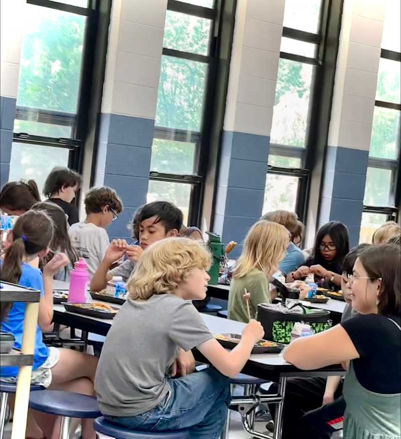 Several elementary aged students are pictured eating lunch and chatting in a school cafeteria with several windows