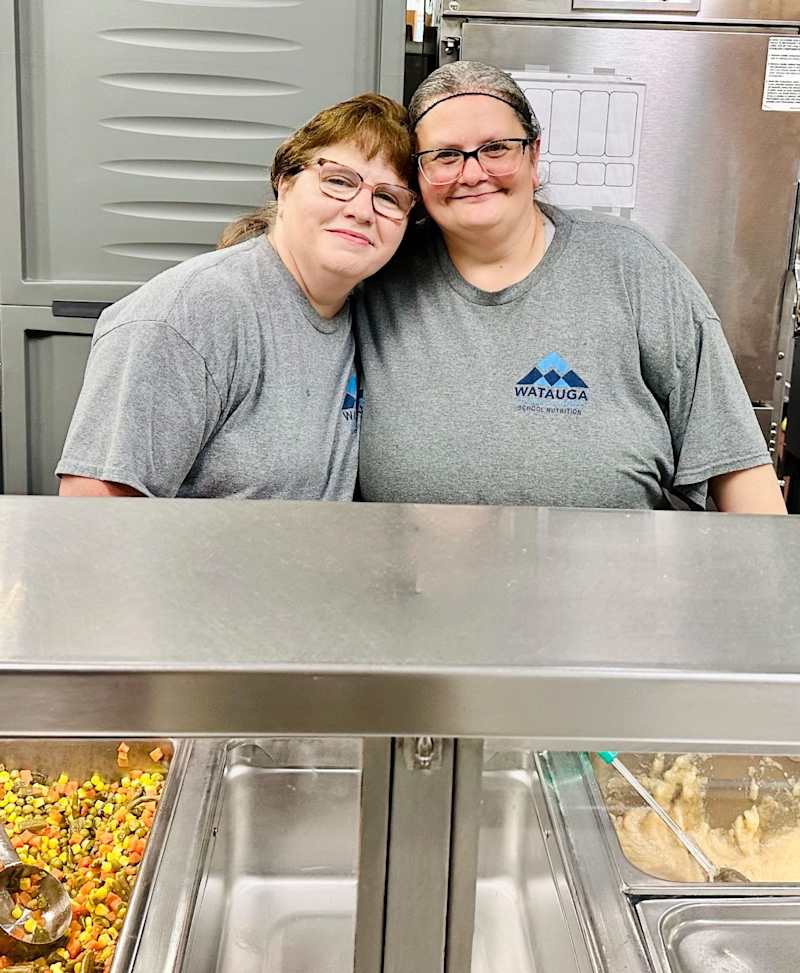 Two adult women dressed in matching grey t shirts with their hair pulled back and glasses on smile for a photo together in a school cafeteria