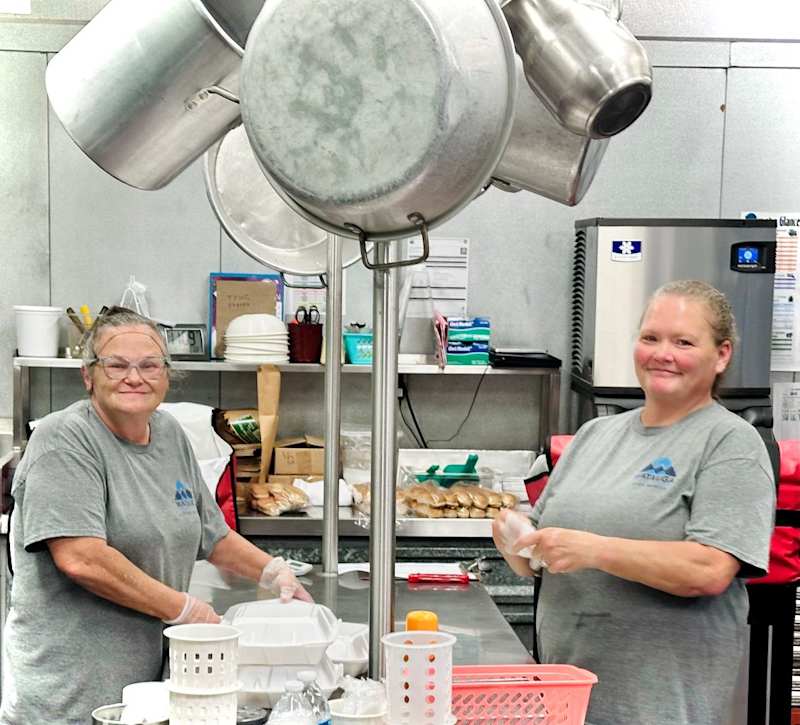 Two women in gray t shirts smile at the camera for a photo while putting on their gloves and packing to go boxes of meals in a school cafeteria
