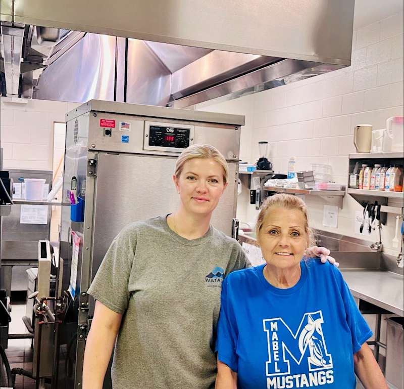 two women one in a gray t shirt and one in a blue t shirt with the school's mascot a mustang and the words "Mabel School" pose for a photo in a school cafeteria.