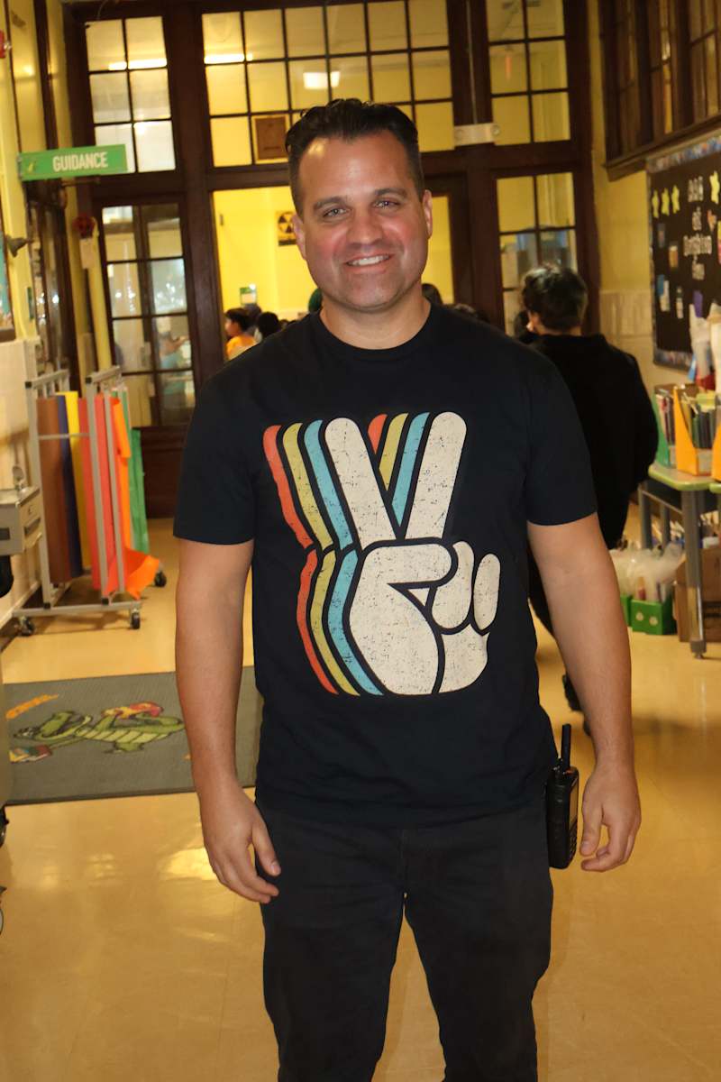 Principal Jason Grimes wears Black T-shhirt with hand holding up Peace Symbol.