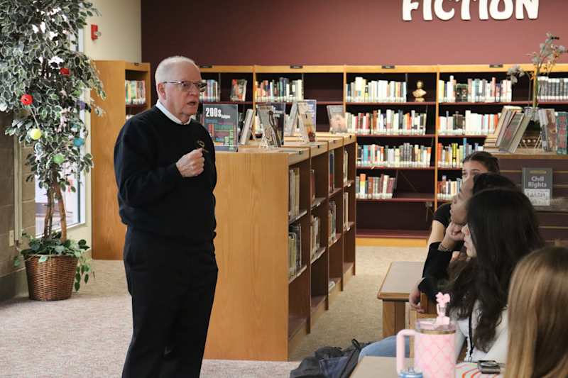 A man in a black veteran sweater stands in front of students seated in a library. He is talking to t…