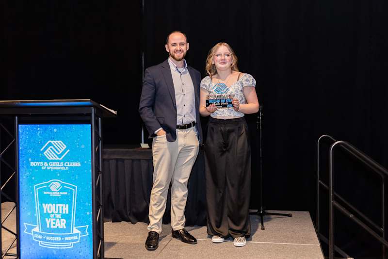 A girl stands on a stage holding a trophy, standing next to a man.