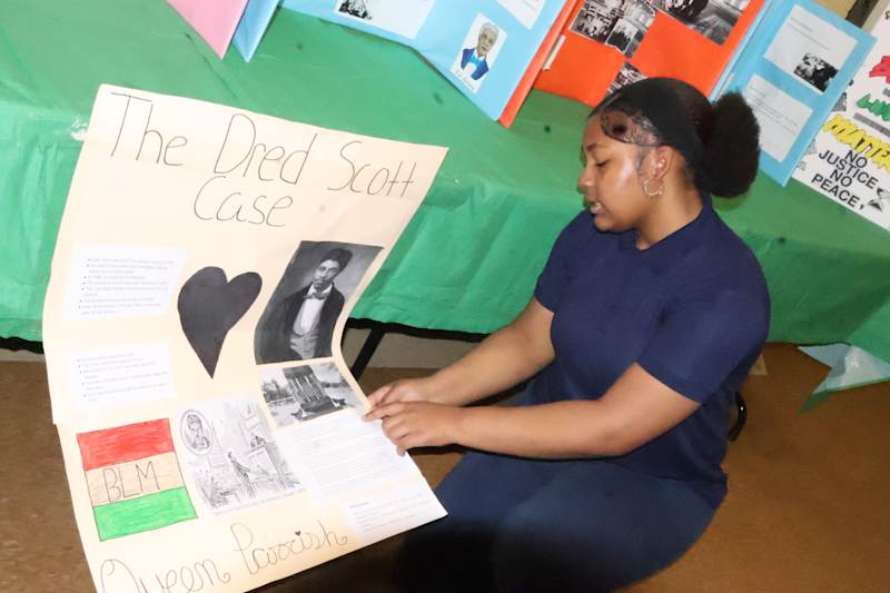 A young person in a navy blue shirt is standing in front of a colorful display board titled "The Dea…