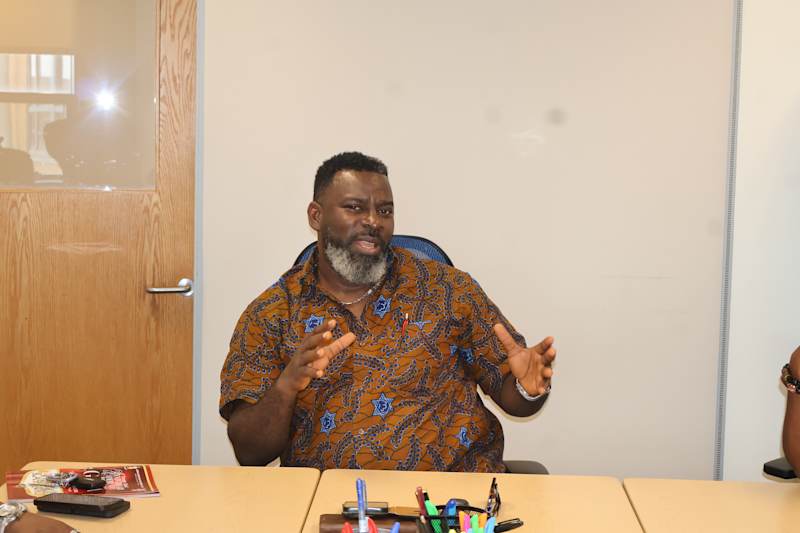 A man with a long beard wearing a colorful, patterned shirt is standing at a table with various item…