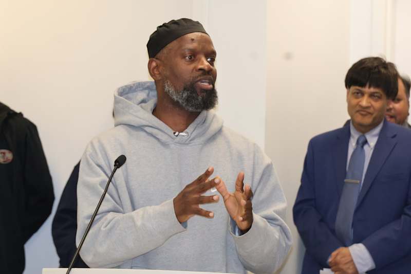 A man with a beard wearing a gray hooded sweatshirt is speaking at a podium, with two other men in s…