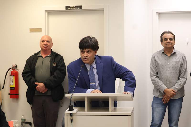 The image shows three men standing in what appears to be an office or workplace setting, with one ma…