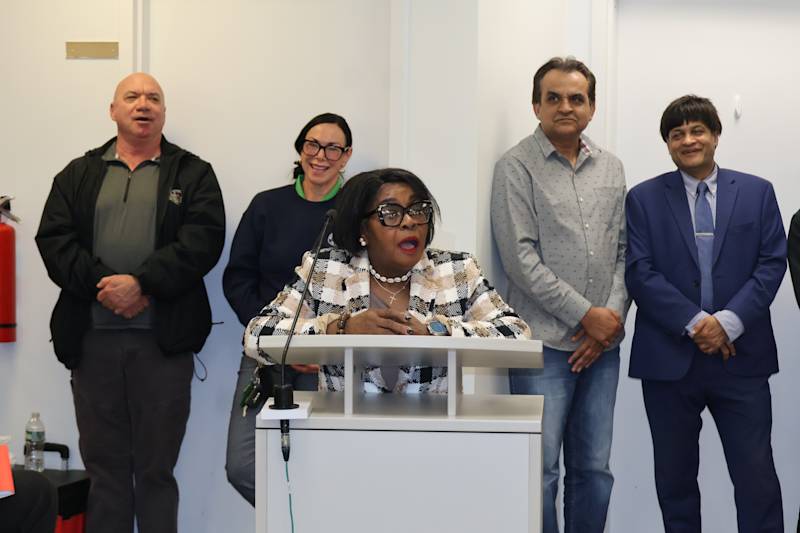 The image shows a group of people, including a woman in the center speaking into a microphone, surro…