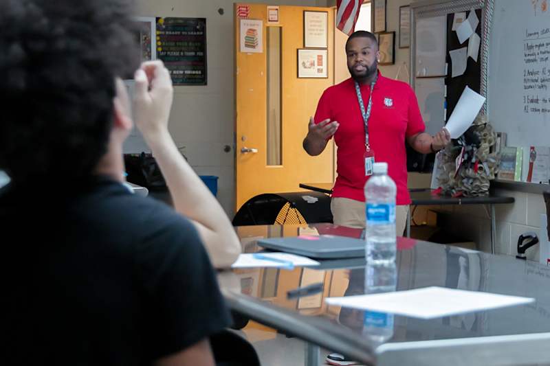 High school culinary teacher standing at the front of a professional instruction kitchen workspace directing students.